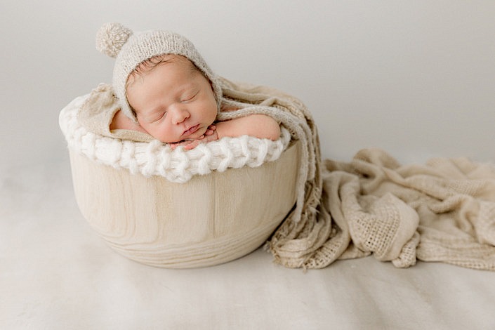 newborn boy wearing a hat while laying with his chin on hands in a light wood bucket with blanket flowing out.
