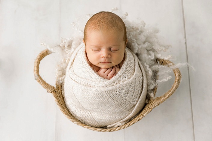 chubby newborn baby boy wrapped up in a textured blanket while sitting inside a wicker basket.