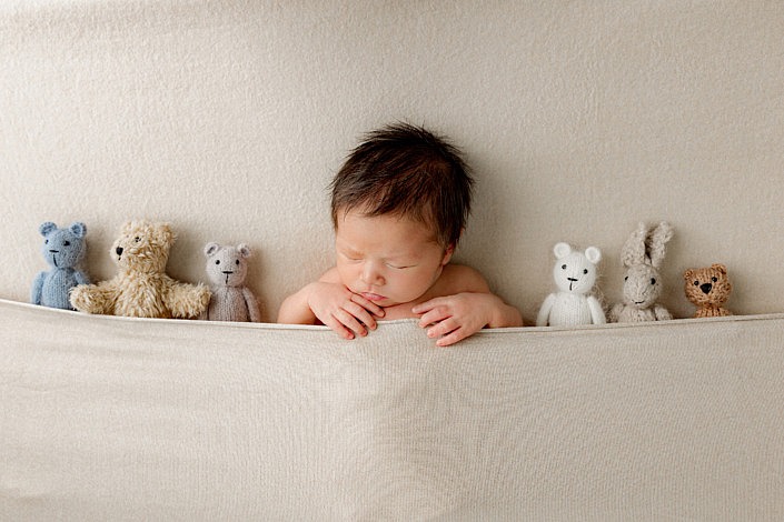 Newborn boy tucked in a tan blanket surrounded by stuffed animals.