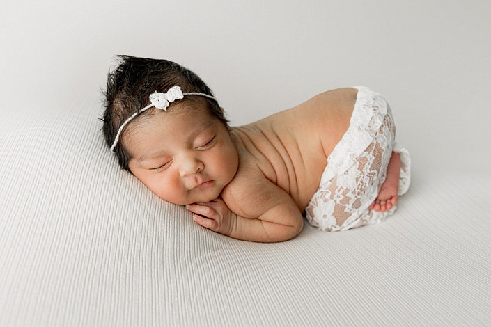 Newborn baby girl on her belly with bum up. She's wearing white lace pants and a white lace bow on her head all against a white background.