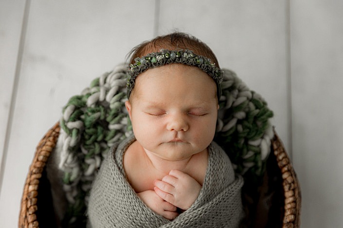 Newborn girl wrapped in a textured green blanket. She is sitting in a basket on a white wood floor.