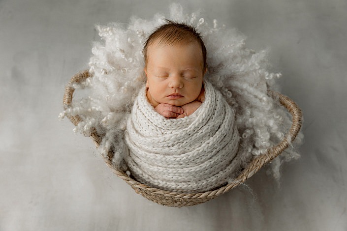 Sweet sleeping newborn baby wrapped in a sweater scarf sitting in a tan basket.