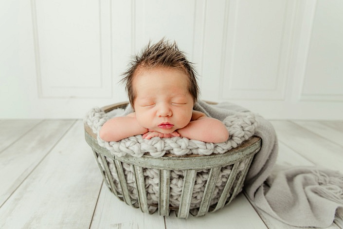 newborn boy showing off his impressive hair while resting on his hands in a grey metal bucket