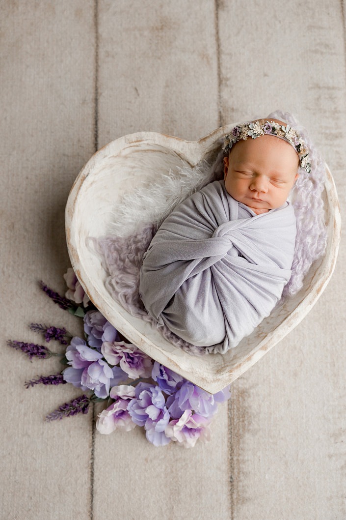 Newborn baby girl wrapped in purple laying in a white heart bowl with purple flowers around the heart