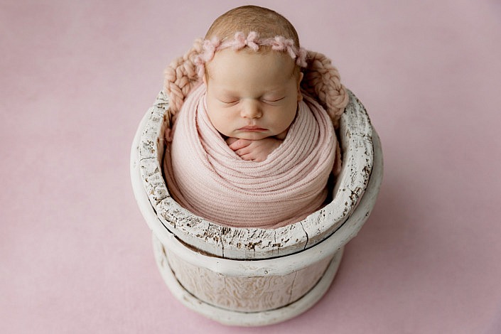 Newborn girl wearing a pink bow headband and pink blanket in a white wooden bucket on a solid pink backdrop.