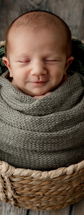 A smiling baby sleeps in a light colored basket on a wood floor. He's wearing a knitted sage green blanket.