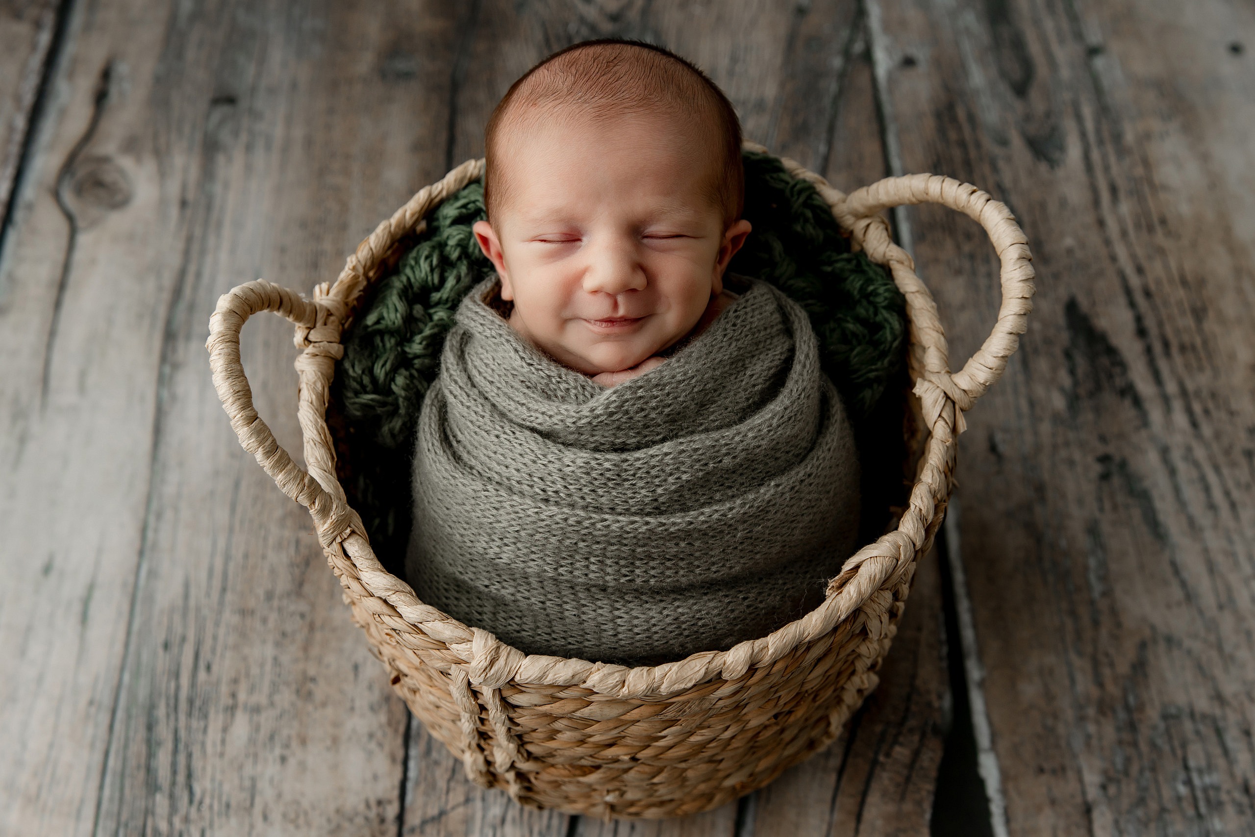 A smiling baby sleeps in a light colored basket on a wood floor. He's wearing a knitted sage green blanket.