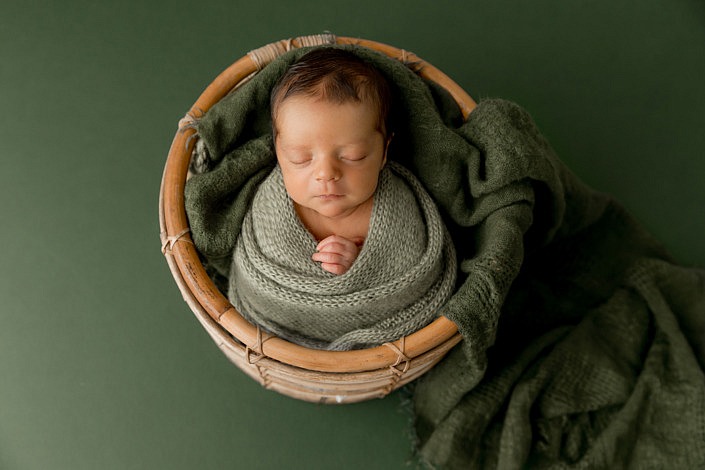 newborn boy wrapped in sage green sitting in a round wooden basket on top of a green background.