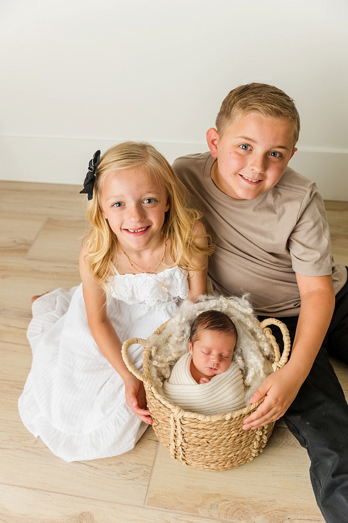 big brother and big sister smile while helping hold their newborn baby brother