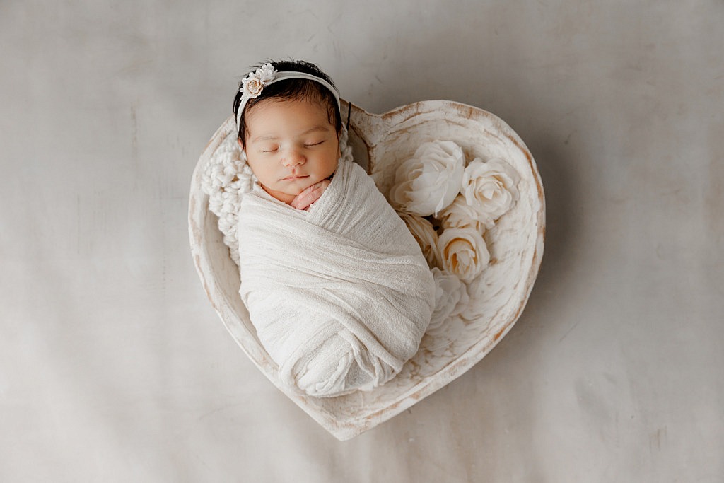 newborn girl wrapped in white and sitting in a heart shaped bowl with white flowers