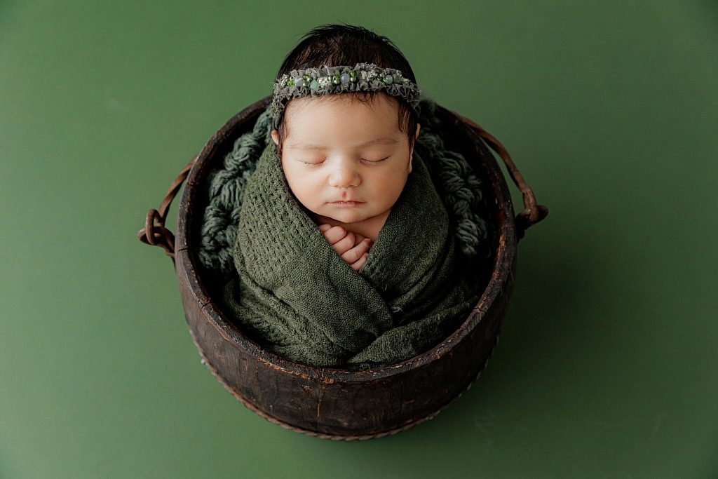 newborn girl wrapped in dark green in a wood bucket