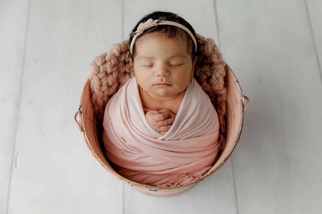 newborn girl wrapped in pink and sitting in a pink bucket on a white wood floor